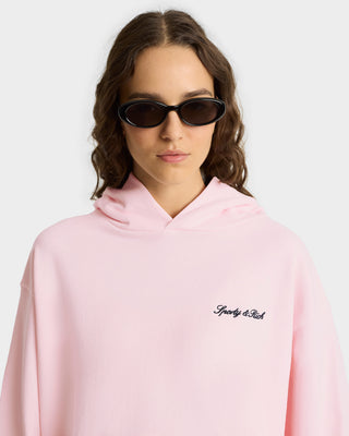 Woman wearing black sunglasses and a light pink Syracuse Embroidered Hoodie - Pink Dolphin, made from 100% USA-grown cotton and featuring an embroidered Sporty & Rich logo on the chest.