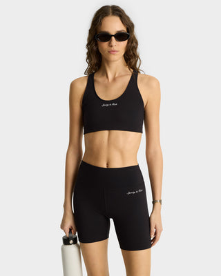 Confident woman in Syracuse Biker Shorts - Black/White and sunglasses holds a water bottle against a plain background—showcasing luxury activewear at its finest.