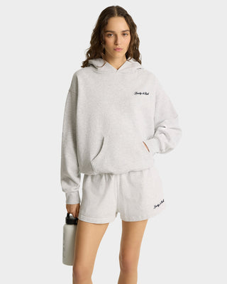 Woman wears the Syracuse Embroidered Hoodie in Heather Gray/Navy, made from USA-grown cotton, paired with shorts, holding a water bottle against a plain background.