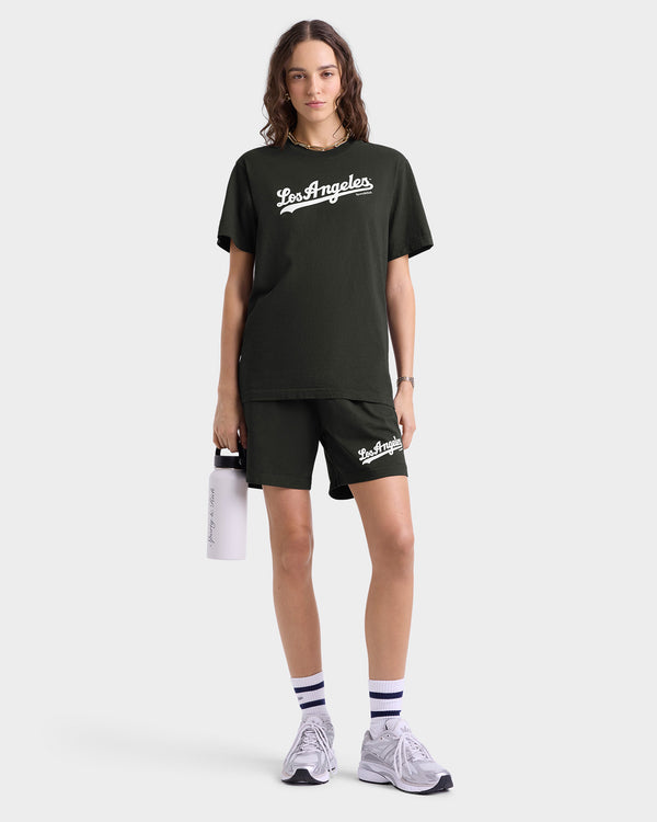 A person in a green Los Angeles shirt and shorts, holding a white water bottle, wearing sneakers and striped socks—proudly showcasing attire made with USA-grown cotton thats shrink-free, including the Dodgers T-Shirt - Faded Black/White.