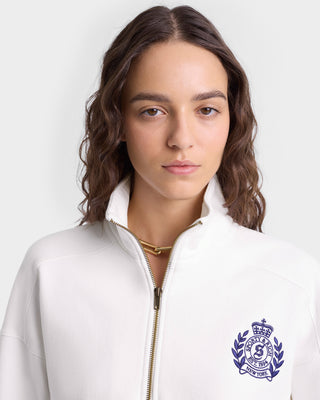 Woman with wavy brown hair wearing the H&W Crest Embroidered Quarter Zip in White/Navy, featuring a 100% cotton fabric and an embroidered crest on the left side.