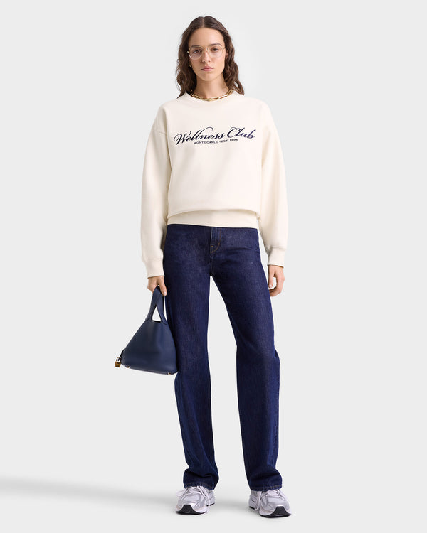 Woman in glasses wears the Wellness Club Embroidered Crewneck - Cream/Navy, made from 100% cotton in Portugal, paired with dark jeans, sneakers, and a navy handbag.
