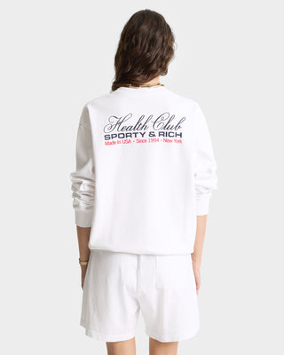 A woman wears the Made In USA Crewneck - White/Navy/Fire Red, a 100% cotton, garment-dyed sweatshirt made in the USA with Health Club Sporty & Rich text on the back.