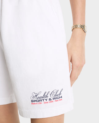 A person wearing Made In USA Gym Short - White/Navy/Fire Red, featuring 100% cotton jersey fabric, Health Club Sporty & Rich text, a drawstring waistband, and a gold wristwatch.