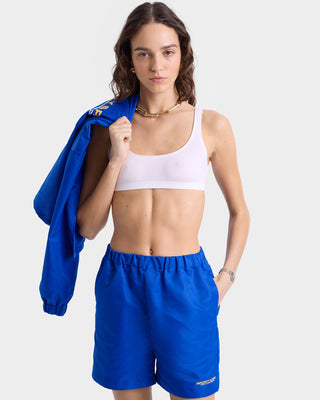 Close up view of model wearing blue nylon shorts with an elastic waistband, a smooth woven finish, and gold 'Sporty & Rich' text near the left hem.