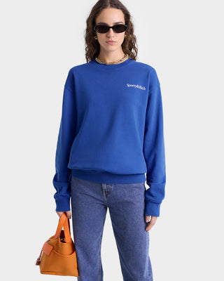 Close up of model wearing royal blue crewneck sweatshirt with white 'Sporty & Rich' text on the left chest, featuring ribbed hem and cuffs.