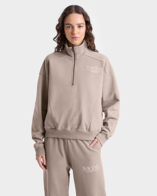 Close up view of model wearing tan sweatpants with an elastic waistband, side pockets, white 'New York Health & Wellness' text on the left thigh, and elasticated cuffs at the ankles, paired with matching quarter zip.