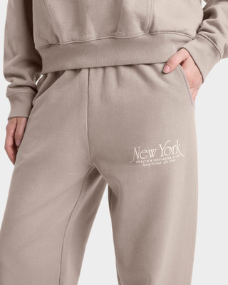 close up view on logo of model wearing tan sweatpants with an elastic waistband, side pockets, white 'New York Health & Wellness' text on the left thigh, and elasticated cuffs at the ankles.