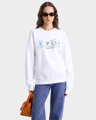 Close up of model wearing white crewneck sweatshirt with a multi-colored 'New York Health & Wellness Club Sporty & Rich' graphic on the chest, featuring ribbed cuffs and hem.