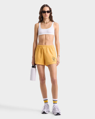 Model wearing gold sweat shorts with an elastic waistband, side pockets, and a black circular 'Sporty & Rich California' graphic near the left hem.