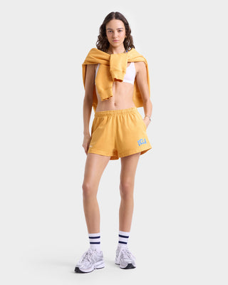 Woman wearing UCLA Ivy Disco Short in gold/blue with an elastic waistband, matching sweatshirt draped over her shoulders, a white crop top, striped socks, and sneakers.