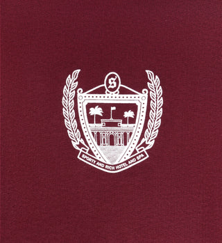 White hotel emblem with palm trees and laurel wreath on maroon fabric; text: Sporty And Rich Hotel And Spa. The Beverly Hills Cropped Hoodie – Merlot is crafted from USA-grown cotton for comfort and timeless style.
